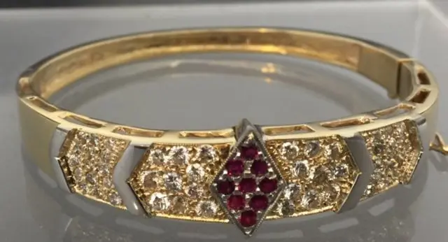 An ornate gold and diamond ring with a ruby center stone, set against a gray background