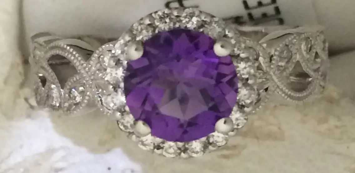 A silver ring with a large oval-shaped purple gemstone in the center, surrounded by smaller clear gemstones