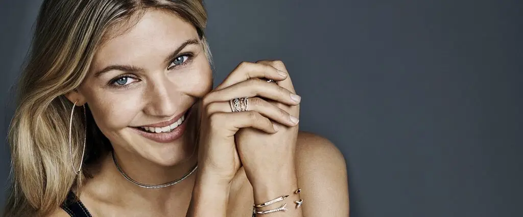A smiling woman with blonde hair wearing various gold-colored jewelry items, including a necklace, bracelet, and ring, against a grey background.