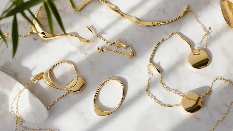 An assortment of gold jewelry including necklaces, bracelets, and rings displayed on a white marble surface with greenery in the background.
